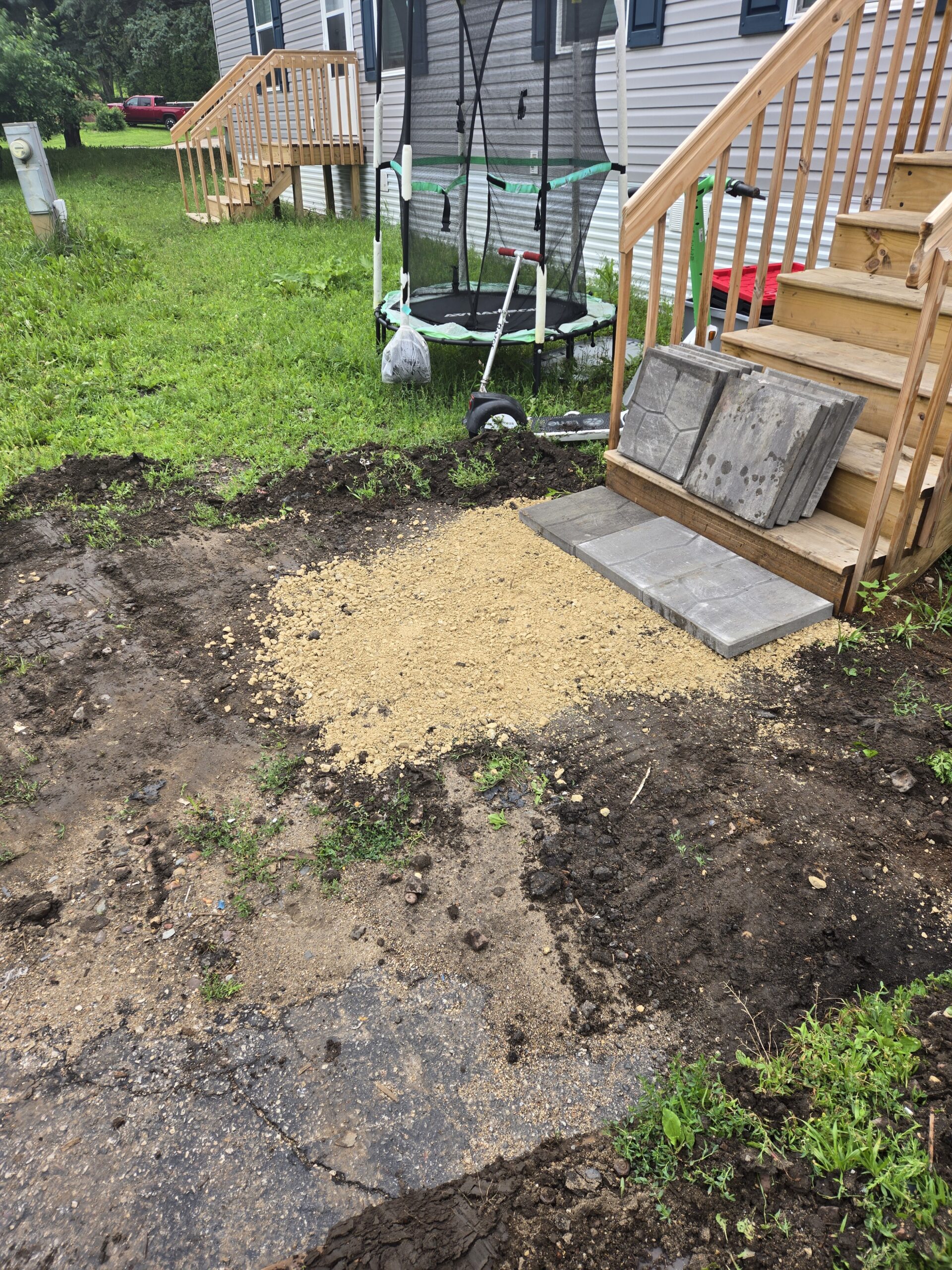 A yard with a small pile of sand next to stairs and a pile of concrete pavers. There is a trampoline in the background.